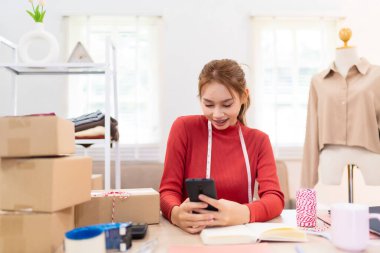 Genç bir kadın masasında oturuyor ve internetteki kıyafet işlerinin siparişlerini yönetmek için telefonunu kullanıyor. Etrafı kutular, defterler ve paketleme malzemeleriyle çevrili.