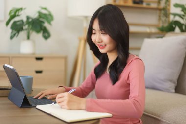 Genç iş kadını tablet üzerinde veri arıyor ve evde çalışırken düşüncelerini kağıda döküyor..