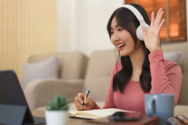 Genç iş kadını iş arkadaşıyla selamlaşıyor ve video görüşmesinde konuşurken kağıda notlar yazıyor.