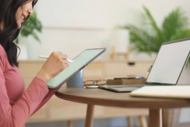 Genç iş kadını tablete iş projesi hakkında notlar yazıyor ve evde dizüstü bilgisayar üzerinde çalışıyor..