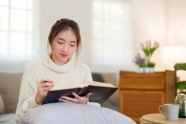 Rahat kanepede oturan genç Asyalı kadınlar rahatlarken ve evde yavaş bir yaşam tarzıyla vakit geçirmek için aktivite yaparken eğitime kitap okumak için kitap açıyorlar..