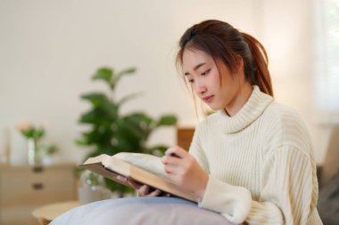 Rahat kanepede oturan genç Asyalı kadınlar rahatlarken ve evde yavaş bir yaşam tarzıyla vakit geçirmek için aktivite yaparken eğitime kitap okumak için kitap açıyorlar..