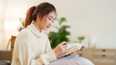 Rahat kanepede oturan genç Asyalı kadınlar rahatlarken ve evde yavaş bir yaşam tarzıyla vakit geçirmek için aktivite yaparken eğitime kitap okumak için kitap açıyorlar..