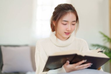 Rahat bir kanepede oturan genç Asyalı kadınlar rahatlarken düşünmek ve öğrenmek için kitap okuyorlar ve evde yavaş yaşam tarzıyla zaman geçirmek için aktivite yapıyorlar..