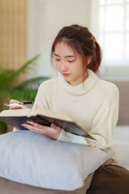 Rahat bir kanepede oturan genç Asyalı kadınlar rahatlarken düşünmek ve öğrenmek için kitap okuyorlar ve evde yavaş yaşam tarzıyla zaman geçirmek için aktivite yapıyorlar..