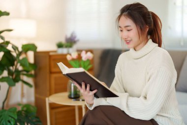 Kazak giyen genç Asyalı kadınlar kitap okuyup edebiyat hikayelerini öğrenirken koltukta rahatlamak ve evde yavaş bir yaşam tarzıyla vakit geçirmek için aktivite yaparken eğleniyorlar..