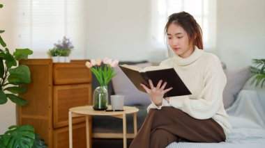 Kazak giyen genç Asyalı kadınlar kitap okuyup edebiyat hikayelerini öğrenirken koltukta rahatlamak ve evde yavaş bir yaşam tarzıyla vakit geçirmek için aktivite yaparken eğleniyorlar..
