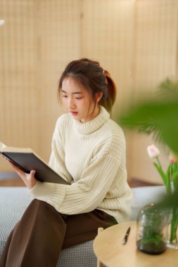 Kazak giyen genç Asyalı kadınlar kitap okuyup edebiyat hikayelerini öğrenirken koltukta rahatlamak ve evde yavaş bir yaşam tarzıyla vakit geçirmek için aktivite yaparken eğleniyorlar..