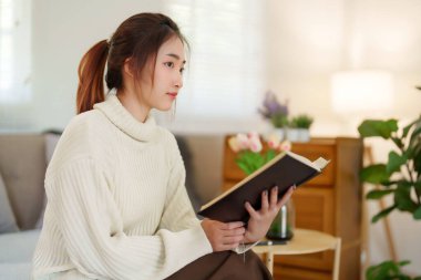Süveter giyen genç Asyalı kadınlar kitap okurken ve edebiyat hikayelerini öğrenirken kanepede oturup dinlenirken ve evde yavaş yaşam tarzıyla zaman geçirmek için aktivite yaparken..