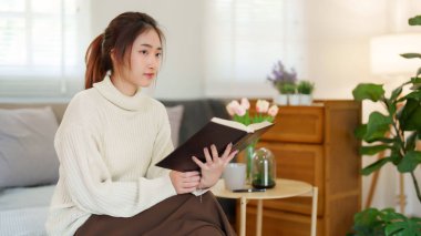 Süveter giyen genç Asyalı kadınlar kitap okurken ve edebiyat hikayelerini öğrenirken kanepede oturup dinlenirken ve evde yavaş yaşam tarzıyla zaman geçirmek için aktivite yaparken..