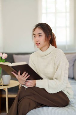 Süveter giyen genç Asyalı kadınlar kitap okurken ve edebiyat hikayelerini öğrenirken kanepede oturup dinlenirken ve evde yavaş yaşam tarzıyla zaman geçirmek için aktivite yaparken..
