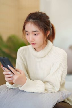 Sörf yapmaktan zevk alan Asyalı genç kadınlar rahat bir kanepede rahatlamak ve evde yavaş bir yaşam tarzıyla vakit geçirmek için etkinlik yaparken akıllı telefonlarıyla sörf yapıyorlar..