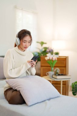 Kazaklı genç Asyalı kadınlar kulaklık takıyor ve akıllı telefondan müzik dinleyip eğlenirken evde yavaş bir yaşam tarzıyla vakit geçirmek için etkinlik yapıyorlar..