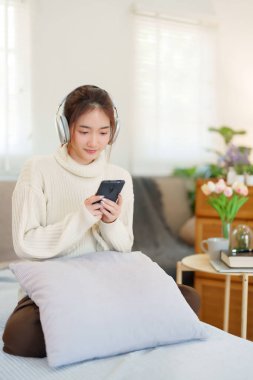 Kazaklı genç Asyalı kadınlar kulaklık takıyor ve akıllı telefondan müzik dinleyip eğlenirken evde yavaş bir yaşam tarzıyla vakit geçirmek için etkinlik yapıyorlar..