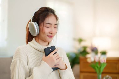 Müzik dinlemek ve nefes almak için kulaklık takan Asyalı genç kadınlar rahatlamak için rahat koltukta otururken ve evde yavaş yaşam tarzıyla zaman geçirmek için etkinlik yaparken..