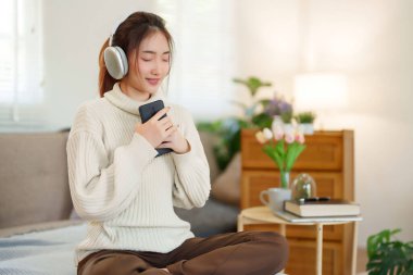 Müzik dinlemek ve nefes almak için kulaklık takan Asyalı genç kadınlar rahatlamak için rahat koltukta otururken ve evde yavaş yaşam tarzıyla zaman geçirmek için etkinlik yaparken..