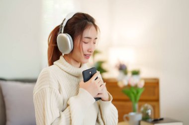 Müzik dinlemek ve nefes almak için kulaklık takan Asyalı genç kadınlar rahatlamak için rahat koltukta otururken ve evde yavaş yaşam tarzıyla zaman geçirmek için etkinlik yaparken..