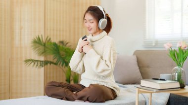 Müzik dinlemek ve nefes almak için kulaklık takan Asyalı genç kadınlar rahatlamak için rahat koltukta otururken ve evde yavaş yaşam tarzıyla zaman geçirmek için etkinlik yaparken..