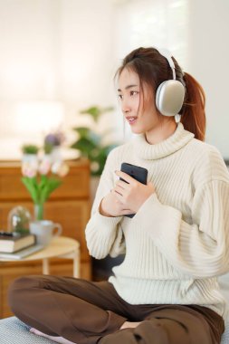 Kazaklı genç Asyalı kadınlar akıllı telefon tutup kulaklık takarak evde yavaş bir yaşam tarzıyla vakit geçirmek için dinlenirken mutlulukla müzik dinliyorlar..