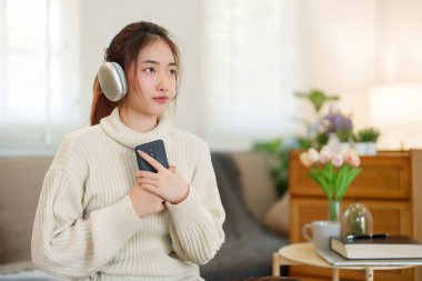Kazaklı genç Asyalı kadınlar akıllı telefon tutup kulaklık takarak evde yavaş bir yaşam tarzıyla vakit geçirmek için dinlenirken mutlulukla müzik dinliyorlar..