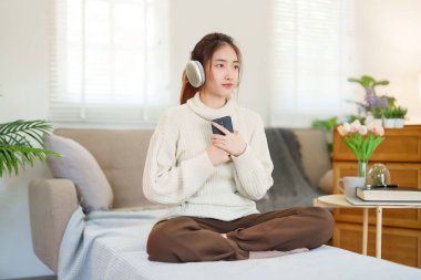 Kazaklı genç Asyalı kadınlar akıllı telefon tutup kulaklık takarak evde yavaş bir yaşam tarzıyla vakit geçirmek için dinlenirken mutlulukla müzik dinliyorlar..