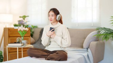 Kazaklı genç Asyalı kadınlar akıllı telefon tutup kulaklık takarak evde yavaş bir yaşam tarzıyla vakit geçirmek için dinlenirken mutlulukla müzik dinliyorlar..