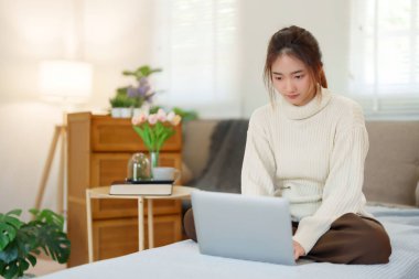 Kanepede oturan Asyalı genç kadınlar ve dizüstü bilgisayarlarıyla sosyal medyada sohbetin keyfini çıkarırken evde yavaş bir yaşam tarzıyla vakit geçirmek için etkinlik yapıyorlar..