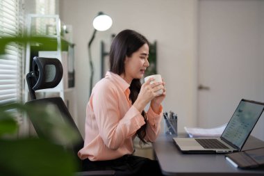 Genç bir kadın masasında oturuyor, elinde bir bardak tutuyor ve dizüstü bilgisayarına bakarken gülümsüyor. O rahat ve modern bir ofis ortamında çalışıyor, işi sırasında bir moladan zevk alıyor.