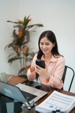 Genç bir kadın masasında oturuyor, dizüstü bilgisayarı üzerinde çalışırken telefonuna bakarken gülümsüyor. Parlak ve rahat bir çalışma alanında.