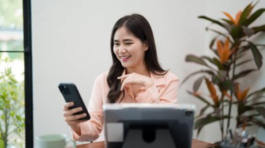 Genç bir kadın masasında oturuyor, dizüstü bilgisayarı üzerinde çalışırken telefonuna bakarken gülümsüyor. Ofis malzemeleri ve yeşilliklerle çevrili, parlak ve rahat bir çalışma alanında..