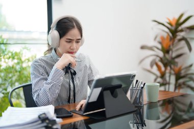 Genç bir kadın masasında oturuyor, kulaklık takıyor ve tablet üzerinde çalışırken odaklanmış görünüyor. Modern, parlak bir ofis ortamında konsantre olmuş gibi görünüyor..