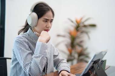 Genç bir kadın masasında oturuyor, kulaklık takıyor ve tablet üzerinde çalışırken odaklanmış görünüyor. Modern, parlak bir ofis ortamında konsantre olmuş gibi görünüyor..