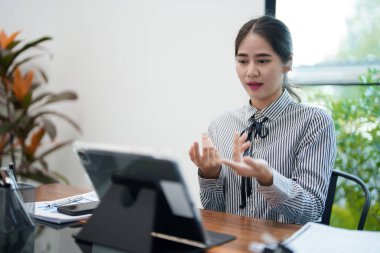 Genç bir kadın masasında oturuyor, tabletindeki bir video görüşmesi sırasında konuşuyor. O, parlak ve modern bir ofiste çalışırken kulaklık takıyor ve elleriyle işaret ediyor..