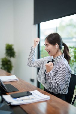 Genç bir kadın masasında oturmuş kulaklık takarken heyecanla yumruklarını kaldırıyor. Sanal bir toplantıda bir başarı ya da olumlu haberi kutluyor gibi görünüyor.