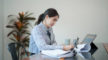 Genç bir kadın masasında oturuyor, belgeleri incelerken gülümsüyor. Odaklanmış ve memnun görünüyor. Parlak, modern bir ofis ortamında çalışıyor.