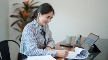 Genç bir kadın masasında oturuyor, belgeleri incelerken gülümsüyor. Odaklanmış ve memnun görünüyor. Parlak, modern bir ofis ortamında çalışıyor.