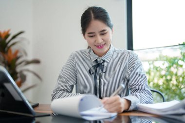 Genç bir kadın masasında oturuyor, belgeleri incelerken gülümsüyor. Odaklanmış ve memnun görünüyor. Parlak, modern bir ofis ortamında çalışıyor.