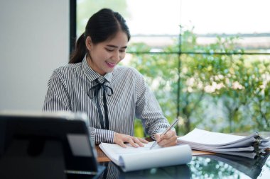 Genç bir kadın masasında oturuyor, belgeleri incelerken gülümsüyor. Odaklanmış ve memnun görünüyor. Parlak, modern bir ofis ortamında çalışıyor.