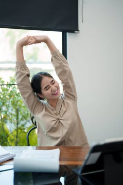 Genç bir kadın masasında oturuyor, kollarını başının üstüne uzatıyor, işe ara veriyor. Parlak, modern bir ofis ortamında rahat görünüyor.