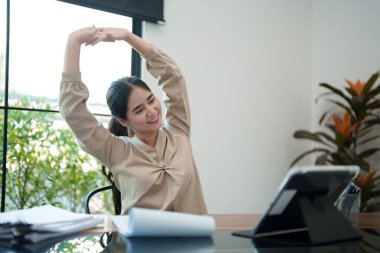 Genç bir kadın masasında oturuyor, kollarını başının üstüne uzatıyor, işe ara veriyor. Parlak, modern bir ofis ortamında rahat görünüyor.