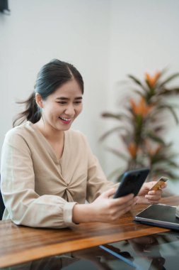 Genç bir kadın masasında oturuyor, telefonu ve kredi kartını tutarken gülümsüyor, muhtemelen internetten alışveriş yapıyor. Modern, parlak bir ofis ortamında.