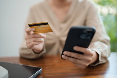 Genç bir kadın masasında oturuyor, telefonu ve kredi kartını tutarken gülümsüyor, muhtemelen internetten alışveriş yapıyor. Modern, parlak bir ofis ortamında.