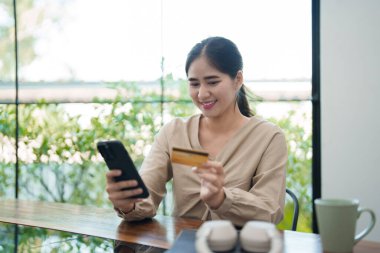 Genç bir kadın masasında oturuyor, telefonu ve kredi kartını tutarken gülümsüyor, muhtemelen internetten alışveriş yapıyor. Modern, parlak bir ofis ortamında.