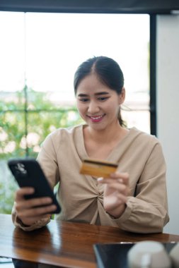 Genç bir kadın masasında oturuyor, telefonu ve kredi kartını tutarken gülümsüyor, muhtemelen internetten alışveriş yapıyor. Modern, parlak bir ofis ortamında.