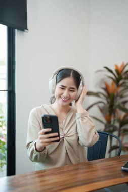 Genç bir kadın masasında oturuyor, kulaklık takıyor ve telefonuna bakarken gülümsüyor. Modern ve parlak bir ofis ortamında çalışan bir sohbet ya da müzikten hoşlanıyor gibi görünüyor..