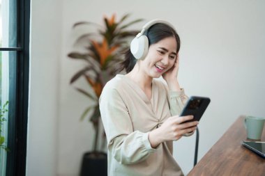 Genç bir kadın masasında oturuyor, kulaklık takıyor ve telefonuna bakarken gülümsüyor. Modern ve parlak bir ofis ortamında çalışan bir sohbet ya da müzikten hoşlanıyor gibi görünüyor..