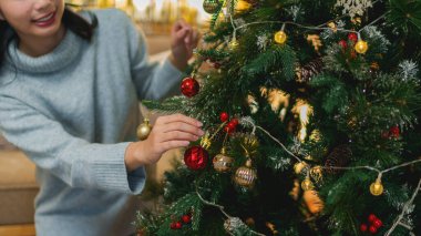 Noel Baba şapkalı genç bir kadın güzel dekore edilmiş bir Noel ağacına süs asmak için uzanır, ışıltılı bir arka planda bayram heyecanıyla gülümser.
