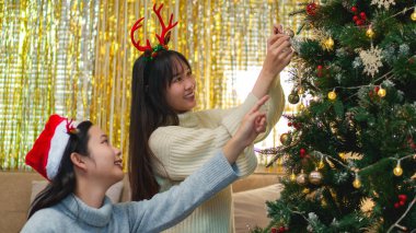 İki arkadaş bir Noel ağacını süslediler, gülümsüyorlar ve Noel Baba şapkaları ve süslemelerle eğleniyorlar..