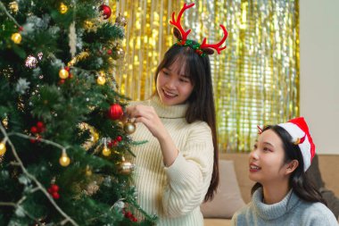 İki arkadaş bir Noel ağacını süslediler, gülümsüyorlar ve Noel Baba şapkaları ve süslemelerle eğleniyorlar..