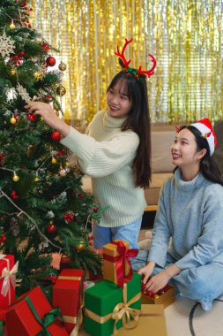 İki arkadaş bir Noel ağacını süslediler, gülümsüyorlar ve Noel Baba şapkaları ve süslemelerle eğleniyorlar..